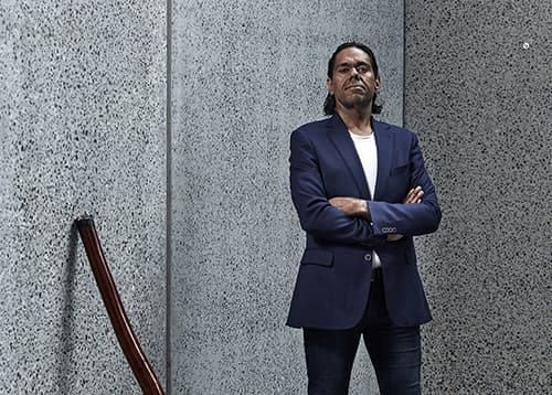 Portrait of William Barton standing with his arms crossed outside of a building with his didgeridoo leaning against the grey speckled concrete walls