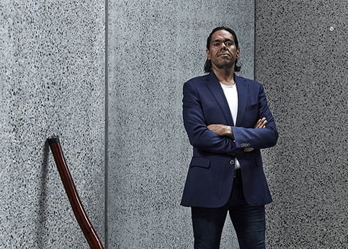 Portrait of William Barton standing with his arms crossed outside of a building with his didgeridoo leaning against the grey speckled concrete walls