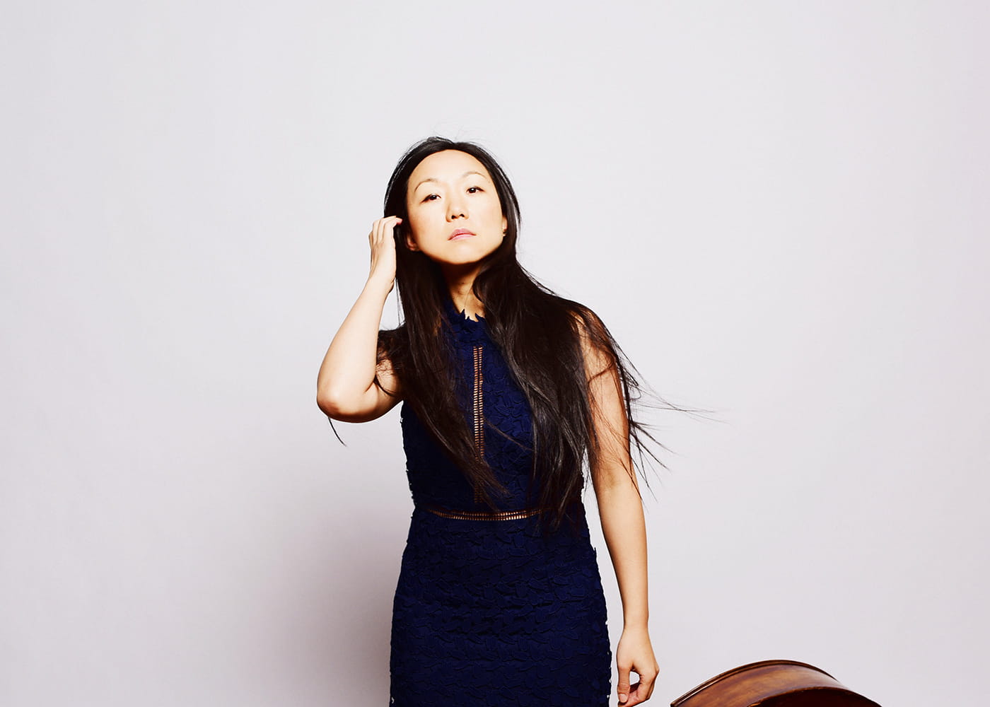 Linda May Han Oh in front of a white background holding her bass in one hand and brushing her hair with the other looking at the camera