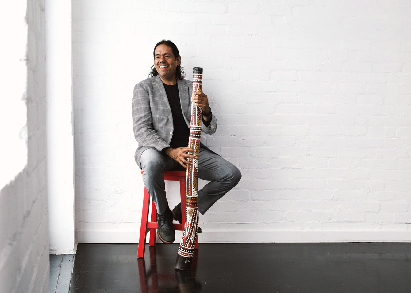 William Barton sitting on a stool in the corner of a sunlit room holding his didgeridoo, looking out of the window